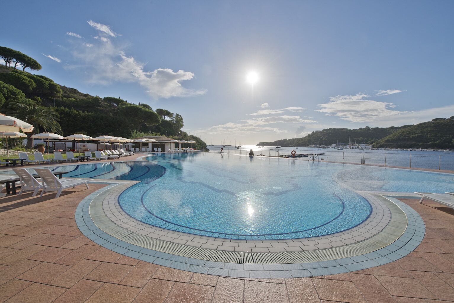 A large outdoor swimming pool with a curved design, surrounded by lounge chairs and umbrellas, with a view of the sea and hills in the background.