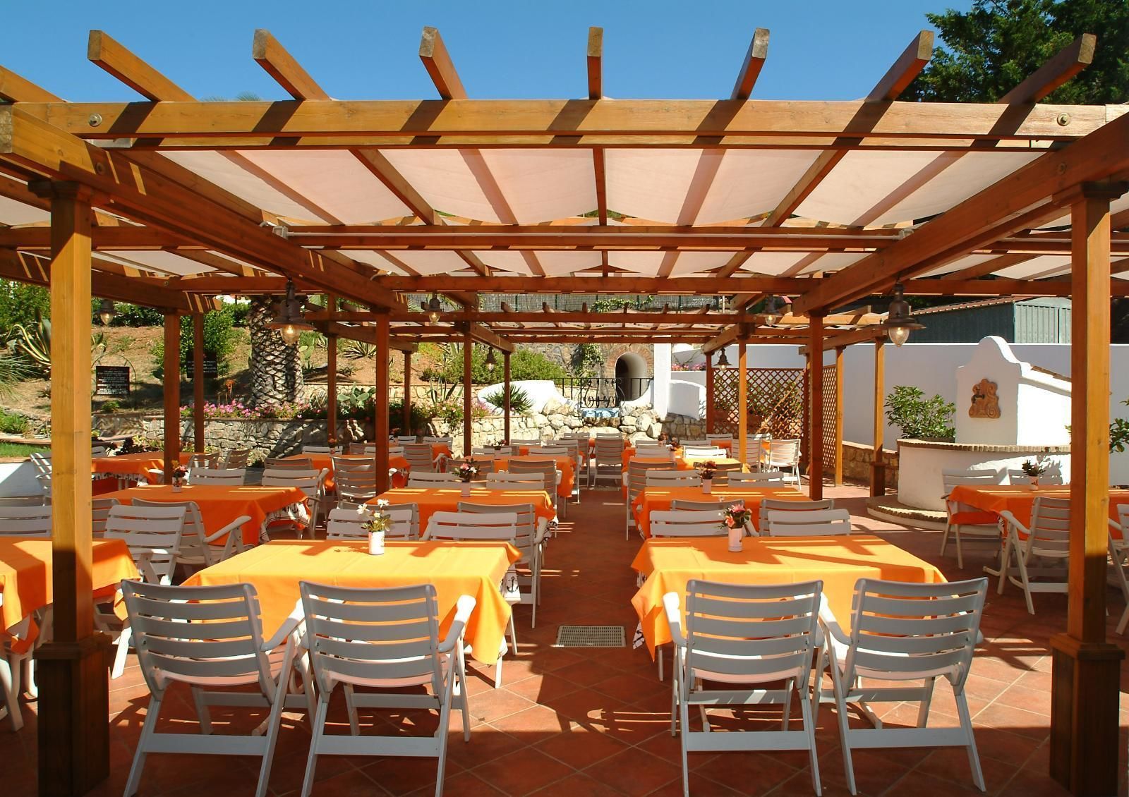 An outdoor dining area with wooden pergolas, orange tablecloths, and white chairs.