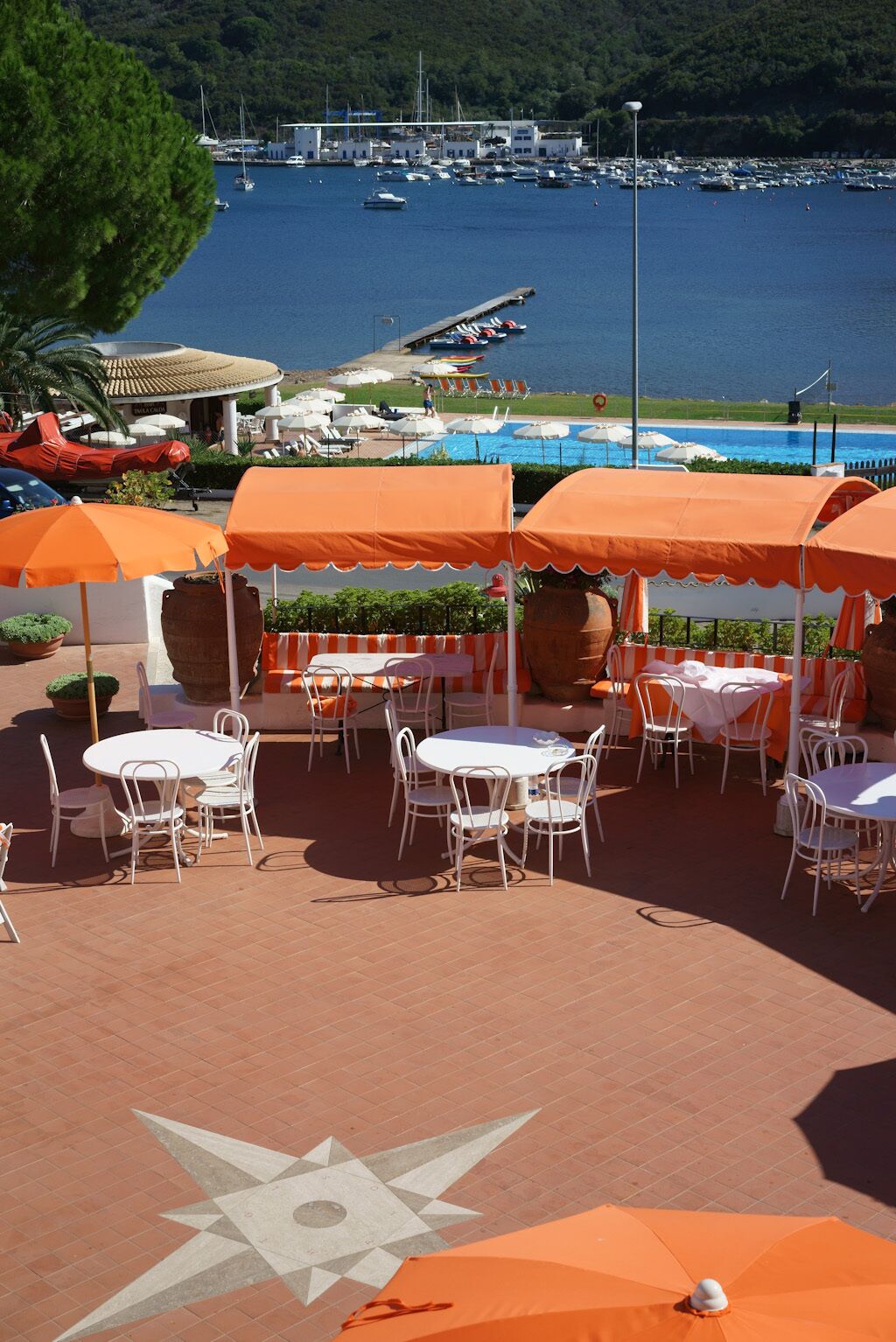 An outdoor dining area with tables and chairs, surrounded by orange umbrellas, overlooking a marina with boats and a swimming pool.