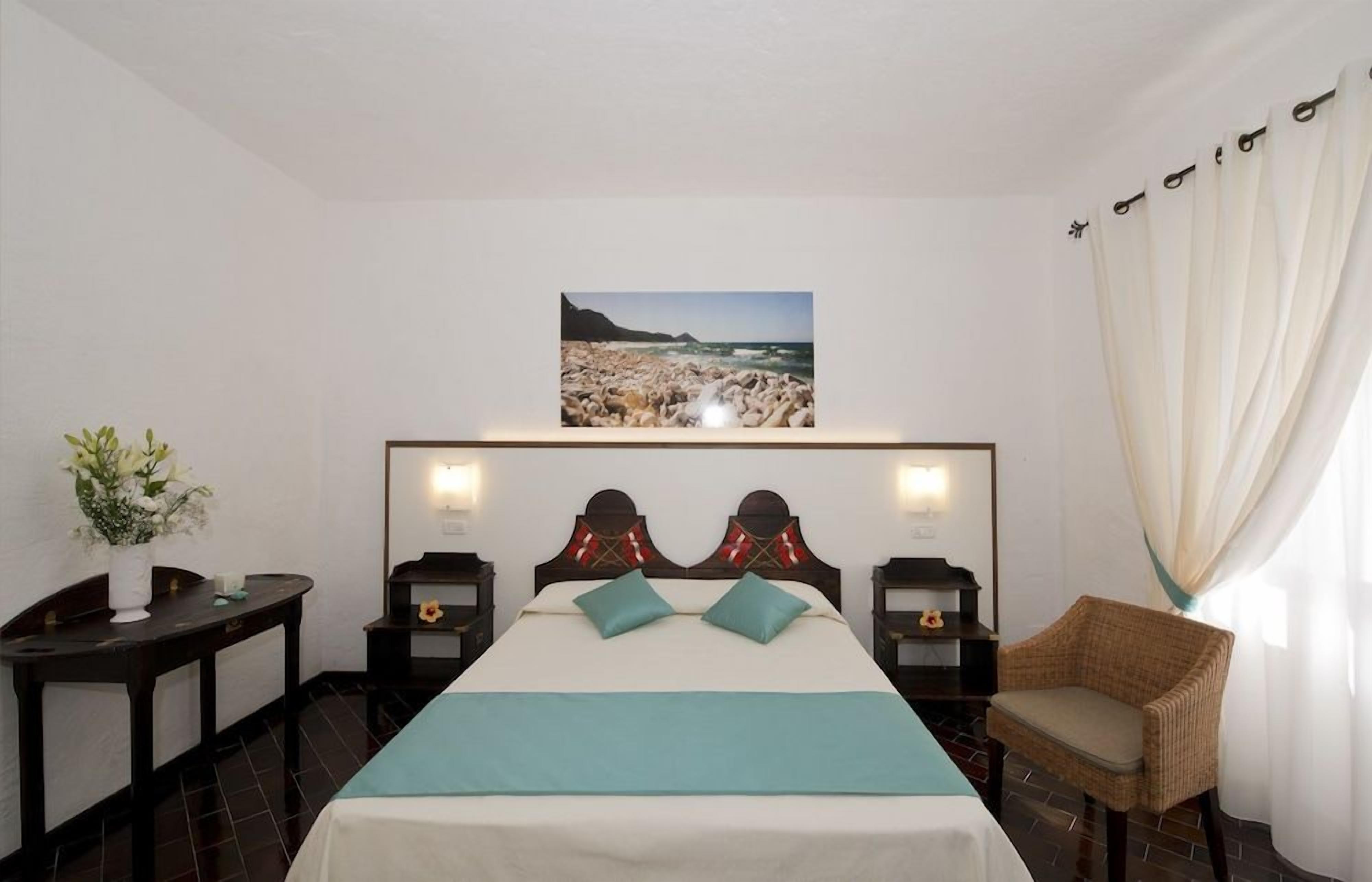 Een hotelkamer met een bed, nachtkastje, stoel, foto van een strand en een gordijn.
