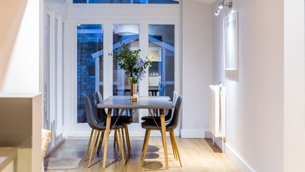 A dining area with a table and chairs, a vase with flowers, and a view through large windows. - The Modern Classic - Contemporary & Elegant 3bdr Home (Cambridge)