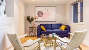 A modern living room with a blue sofa, white wicker chairs, a glass coffee table, and a framed abstract painting. - The Modern Classic - Contemporary & Elegant 3bdr Home (Cambridge)