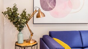 A room with a blue sofa, a small table with a plant and a lamp, and a wall with a framed abstract painting. - The Modern Classic - Contemporary & Elegant 3bdr Home (Cambridge)