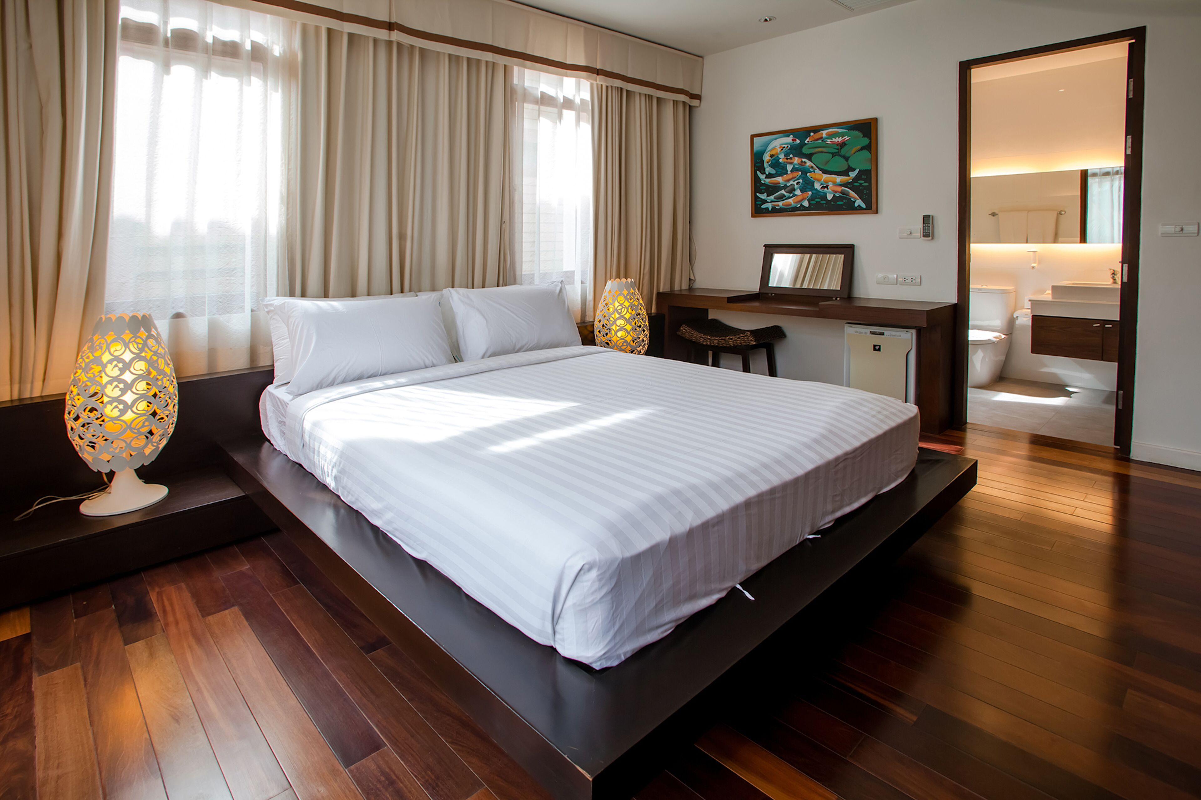 A modern hotel room with a large bed, wooden flooring, a desk with a chair, a small bathroom, and a painting on the wall.