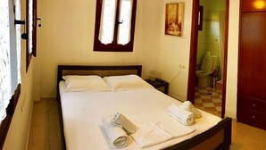 Ein ordentlich bezogenes Bett mit weißen Leinen und zwei zusammengerollten Handtüchern, ein hölzernes Kopfteil, ein Nachttisch mit Lampe und eine Tür zu einem Badezimmer.