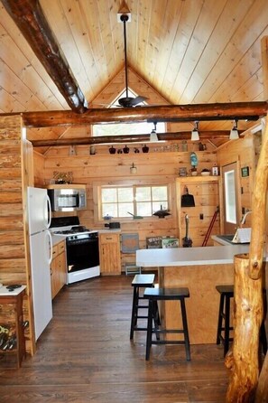 A log cabin with a kitchen featuring a stove, microwave, and refrigerator. There are wooden floors and a wooden ceiling with exposed beams. - Merchant's Log Cabin (Hancock)