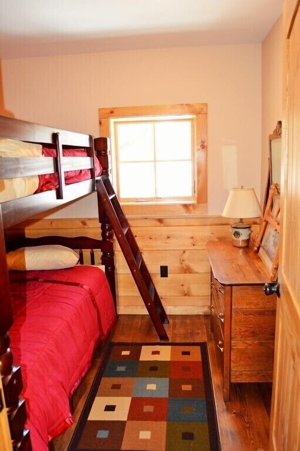 A bunk bed with a red bedspread, a wooden dresser, a lamp, and a window with a view. - Merchant's Log Cabin (Hancock)