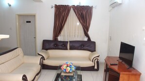 A living room with a beige sofa, a glass coffee table with a floral centerpiece, a TV on a wooden stand, and brown curtains. - Barcelona Apartment (Abuja)