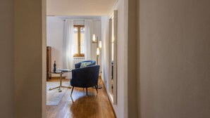 En hall med trägolv, en blå fåtölj, ett litet bord med en flaska och glas, och ett fönster med gardiner.