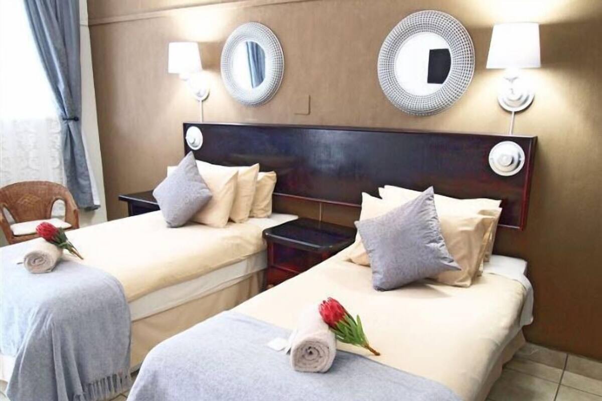 A hotel room with two beds, each with a pillow and a rolled towel. There are two wall-mounted lamps and two mirrors. A chair and a red flower are also visible.