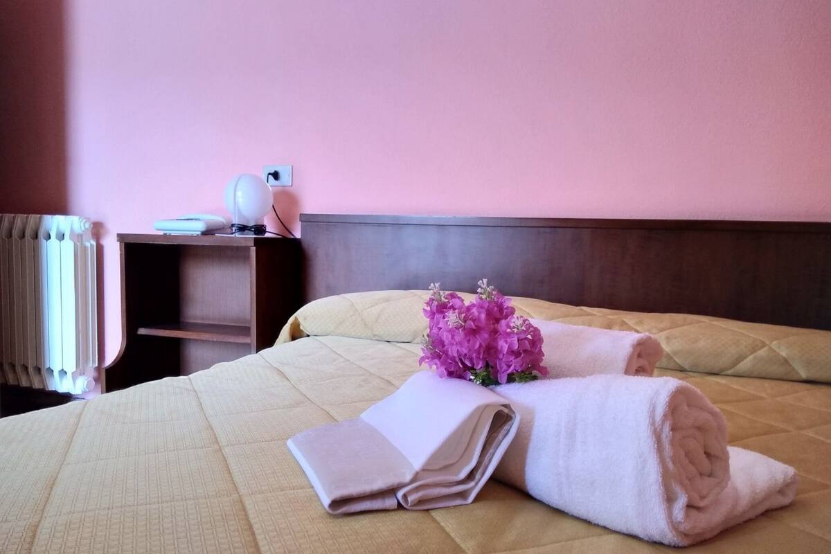 Un lit bien fait, recouvert d’une courtepointe beige, agrémenté d’un bouquet de fleurs roses et de serviettes enroulées, dans une chambre d’hôtel dotée d’une tête de lit en bois et d’une table de chevet avec une lampe.