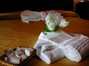 Ein ordentlich bezogenes Bett mit goldfarbener Tagesdecke, zwei gefaltete weiße Handtücher, ein kleiner Korb mit Pflegeprodukten und eine Vase mit weißen Blumen.