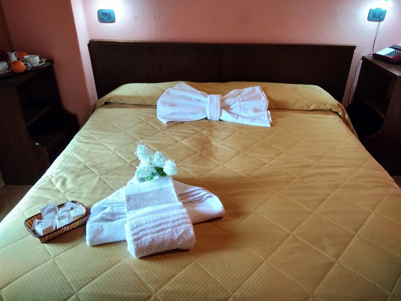 A hotel room with a bed, pillows, a bouquet of flowers, and a basket of toiletries.