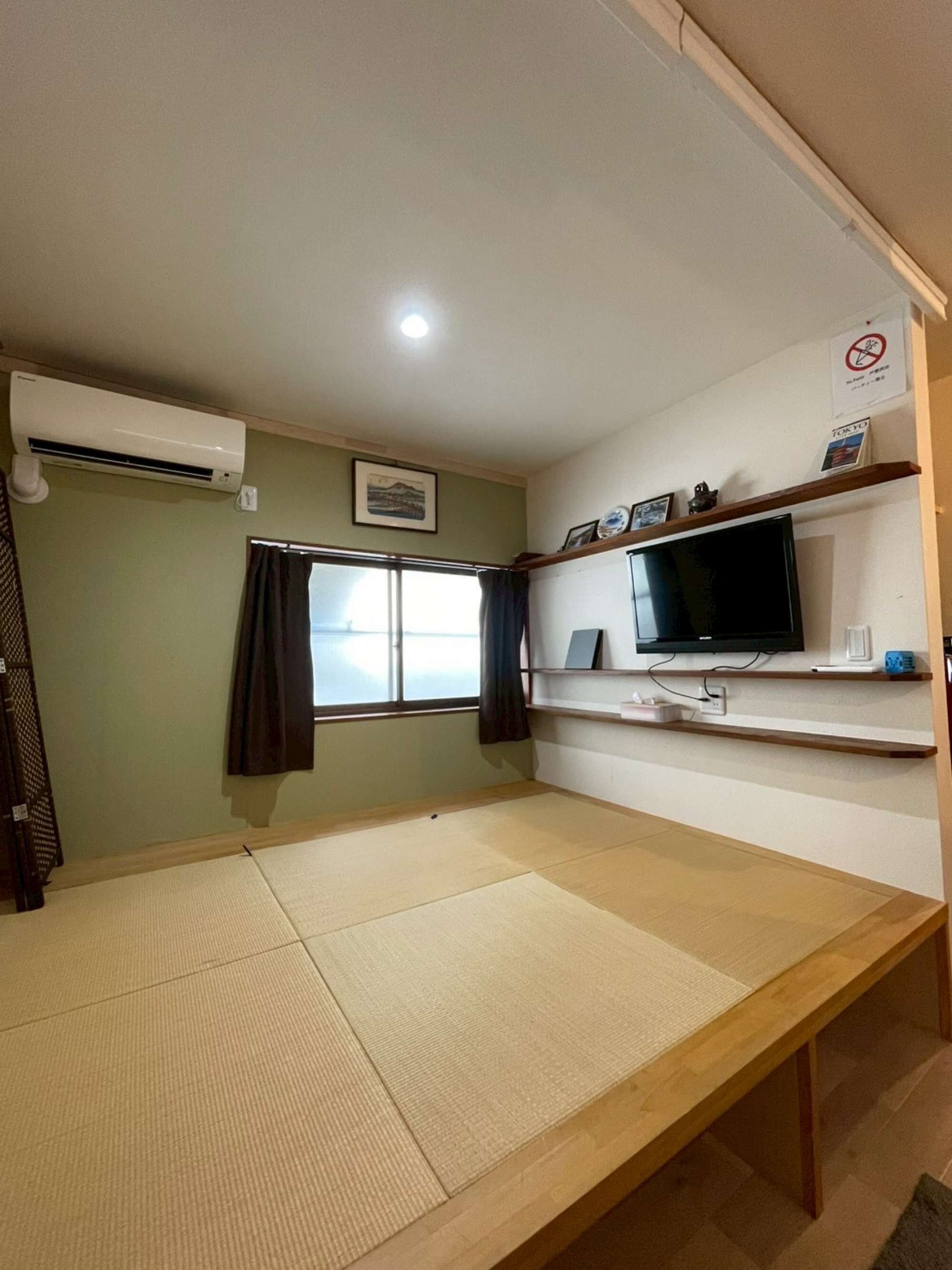 Ein Zimmer mit Tatami-Böden, einem Fernseher, einem Regal mit Dekoration und einem Fenster mit Vorhängen.