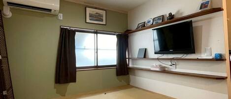 Ein Zimmer mit Tatami-Böden, einem Fernseher, einem Regal mit Dekoration und einem Fenster mit Vorhängen.