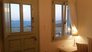 Une chambre avec une porte donnant sur un balcon d’où l’on peut admirer la mer et les montagnes. On y trouve une lampe de chevet et une petite table sur laquelle est posée une autre lampe.