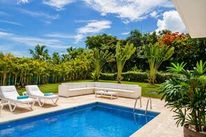 Zona junto a la piscina con un sofá blanco, dos sillones de descanso y una mesita.