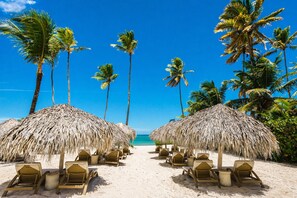 A beach with thatched umbrellas and lounge chairs, palm trees, and a clear blue sky.