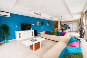 Una sala de estar moderna con una pared azul decorativa, un sofá blanco, cojines de colores, una mesa de centro, un televisor y un ventilador de techo.