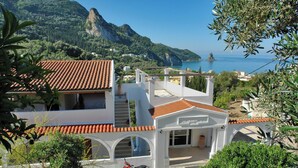 Um edifício costeiro com telhado de telhas vermelhas, fachada branca e vista para o mar e montanhas.