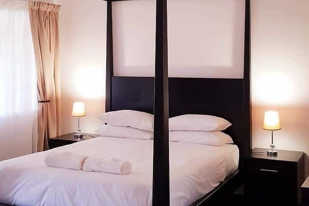 Un lit à baldaquin avec une literie blanche, deux tables de chevet avec des lampes et une grande fenêtre avec des rideaux.