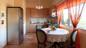 A compact kitchen with wooden cabinets, a refrigerator, a dining table with chairs, and a window with curtains. - PEGGY'S VILLA 50 SQM (Κινέτα)
