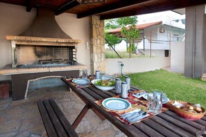 Un área de comedor al aire libre con una parrilla de piedra, una mesa dispuesta con platos, cubiertos y bebidas, y una vista de una zona residencial al fondo.