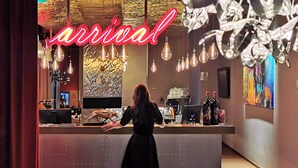 Une femme vêtue d’une robe noire se tient à l’accueil d’un hôtel, où un panneau néon indique « arrivée ». L’intérieur est moderne, avec des luminaires suspendus, et un bar est aménagé avec des bouteilles et des verres.