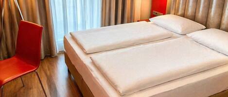 Una habitación de hotel con una cama, una silla roja, una lámpara y una ventana con cortinas.