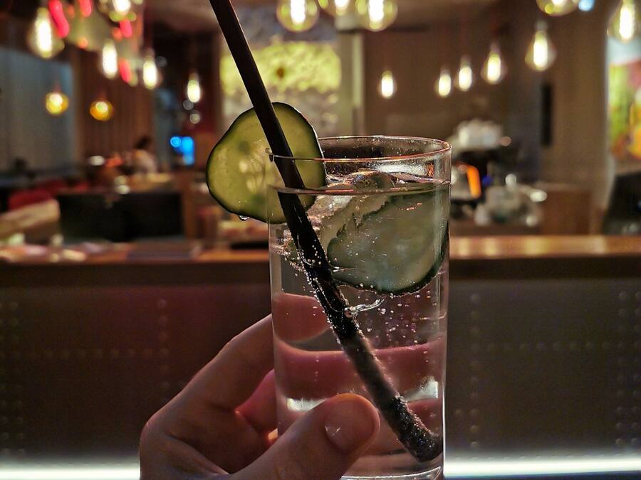 Uma mão segurando um copo com canudo, contendo uma bebida com uma fatia de limão e gelo, em um bar com um letreiro de neon ao fundo.