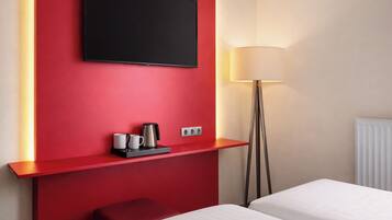 Habitación de hotel con una pared de acento roja, un televisor de pantalla plana sobre un estante con hervidor y tazas, una lámpara de noche y dos camas.