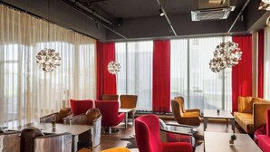 Un hall d’hôtel moderne avec des fauteuils rouges et orange, une table en bois et une grande fenêtre avec des rideaux.