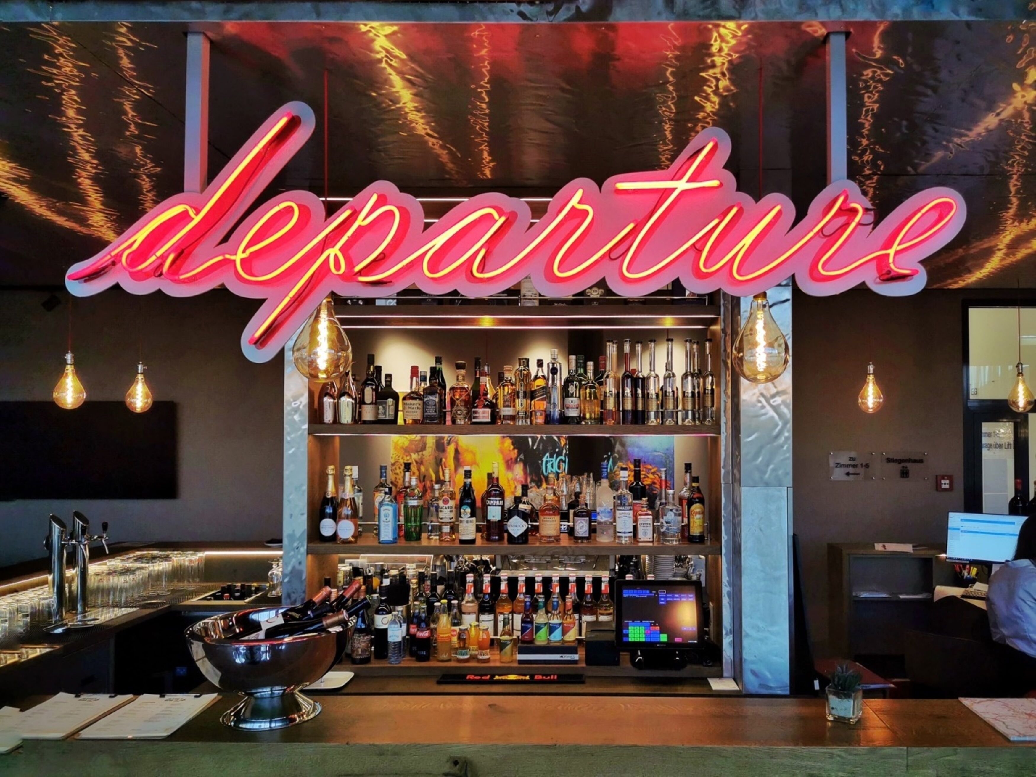 Un coin bar agrémenté d’un panneau néon indiquant le mot « départ » et de diverses bouteilles d’alcool exposées sur des étagères.
