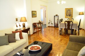 A living room with a sofa, a coffee table with a bowl, a dining table with chairs, and a painting on the wall. - PEDION AREOS PARK 5, sleep 8-12, 4-bedr, 2 bathrooms, Metro, Museum in 150m (Αθήνα)