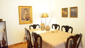 Een eetkamer met een tafel gedekt voor vier personen, omringd door houten stoelen. Twee ingelijste schilderijen hangen aan de muur, er staat een lamp op de tafel en een vaas met takken.