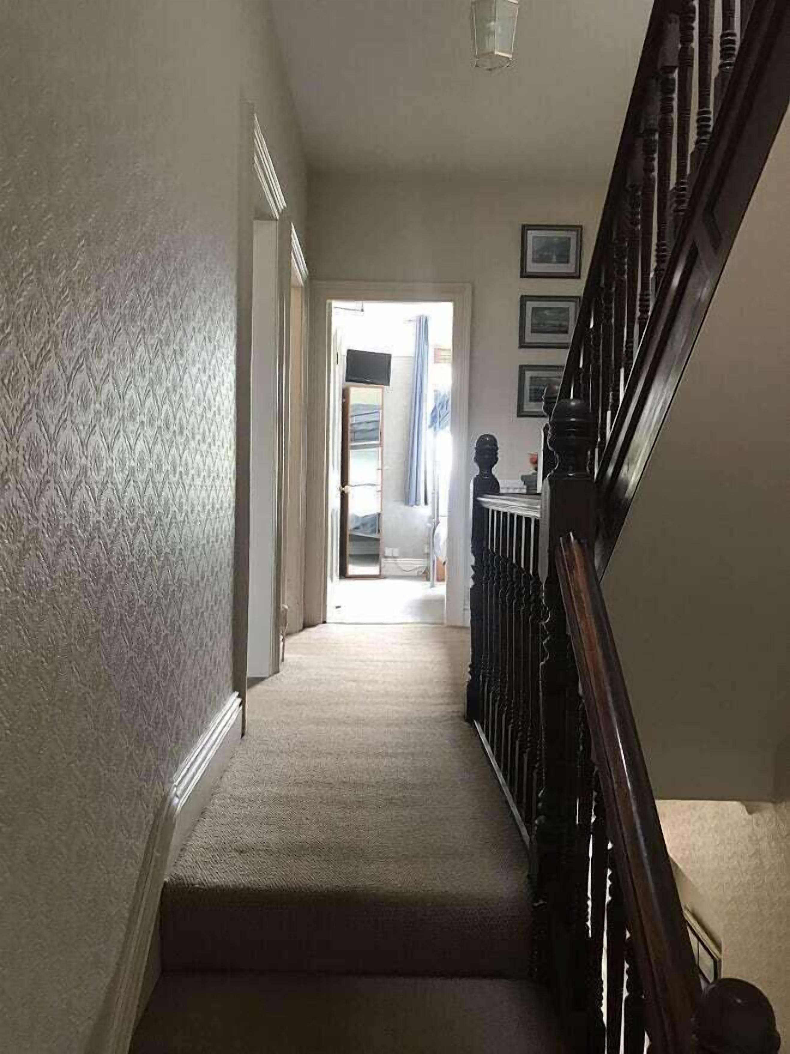 A carpeted hallway with a wooden railing, leading to a room with a bed and a window with curtains.