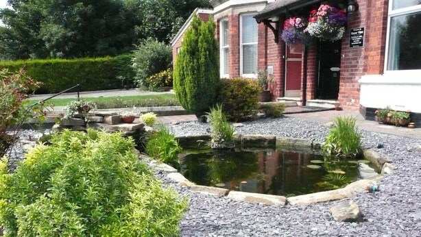 A landscaped garden with a pond, a brick building with a red door, and a sign indicating a view of the sea. - Manor View Guest House (Whitby)