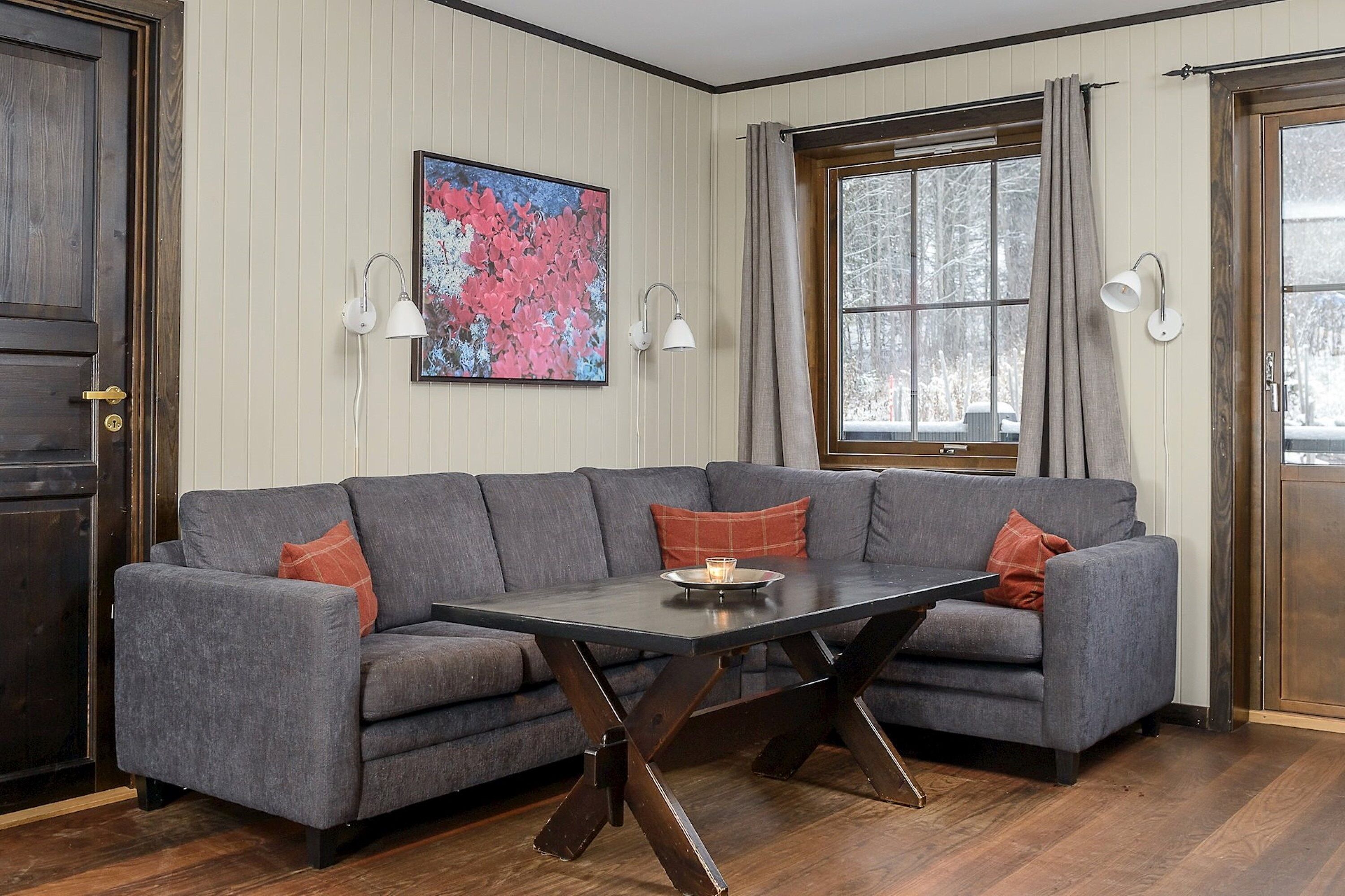 A room with a gray sectional sofa, a dark wooden coffee table, and a large window with curtains.