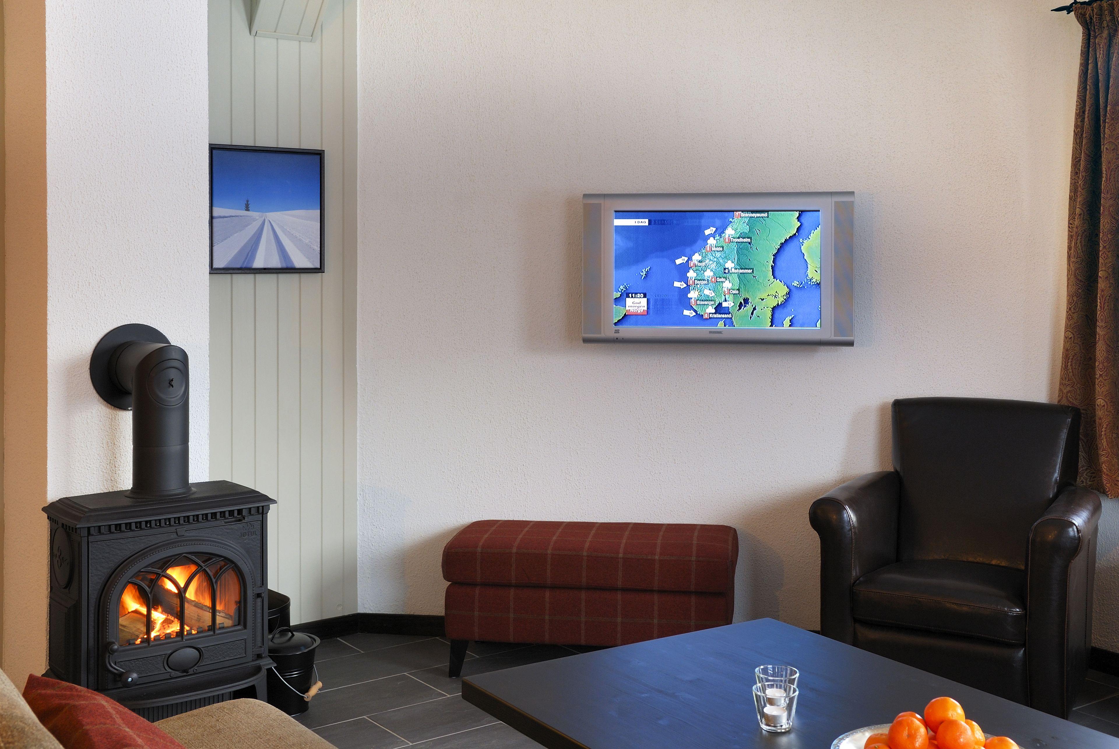 A cozy room with a wood-burning stove, a television mounted on the wall, a red plaid bench, and a black leather chair.