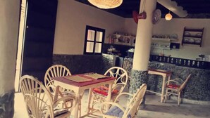 A dining area with white chairs and tables, a stone wall, and a kitchen in the background. - Gîte d'étape Imsker (Marrakech)
