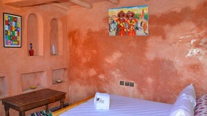 A room with a bed, a wooden table, a colorful painting, and a wall-mounted shelf with a red container. - Gîte d'étape Imsker (Marrakech)