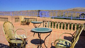 An outdoor seating area with wicker chairs and round tables, a blue railing, and a view of a mountainous landscape. - Gîte d'étape Imsker (Marrakech)