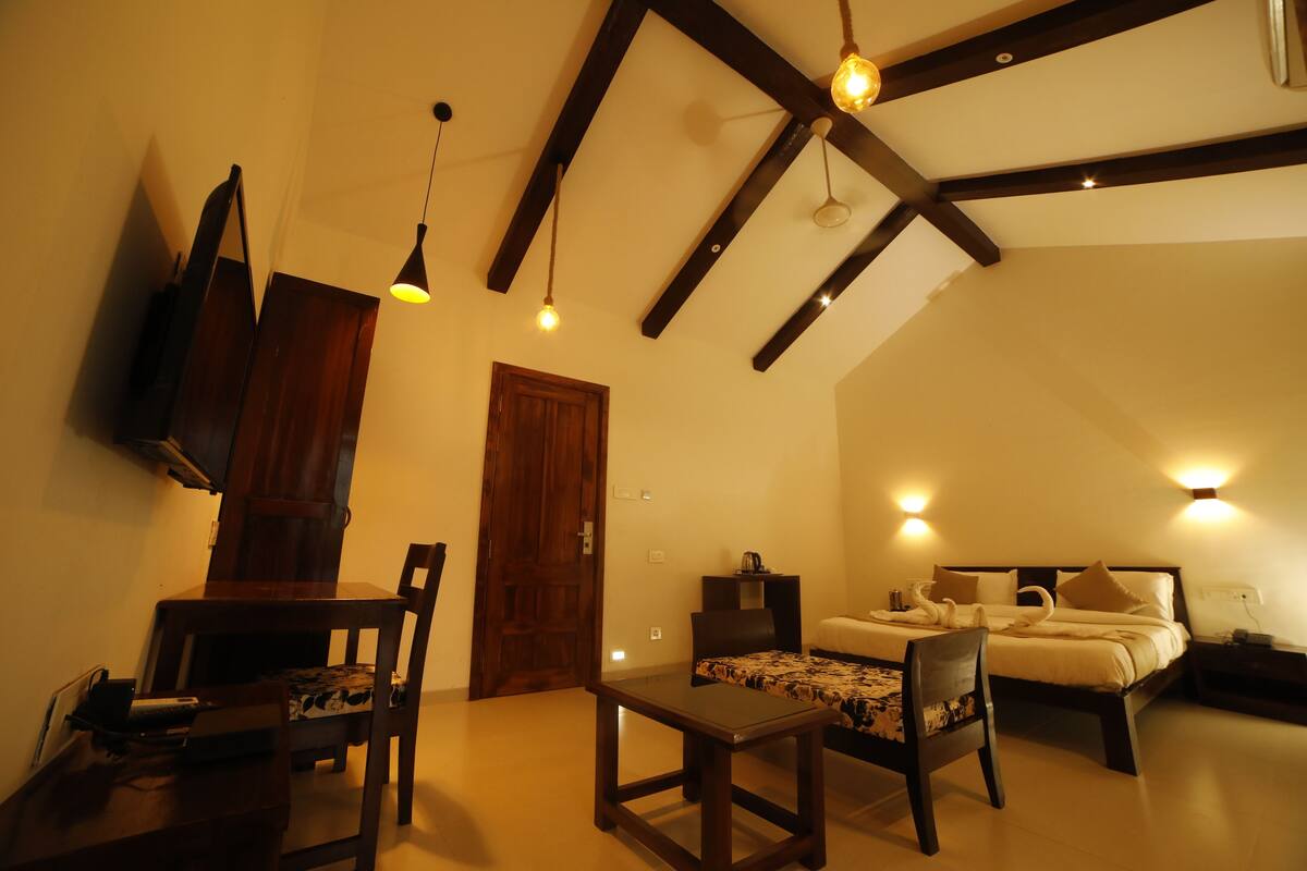 A cozy room with a bed, a small table, and a chair. There is a wooden door and a wall-mounted TV.