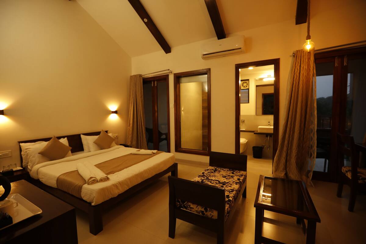 A hotel room with a large bed, a desk, a chair, and a small table. There is a window with curtains, a mirror, and an air conditioning unit on the wall.