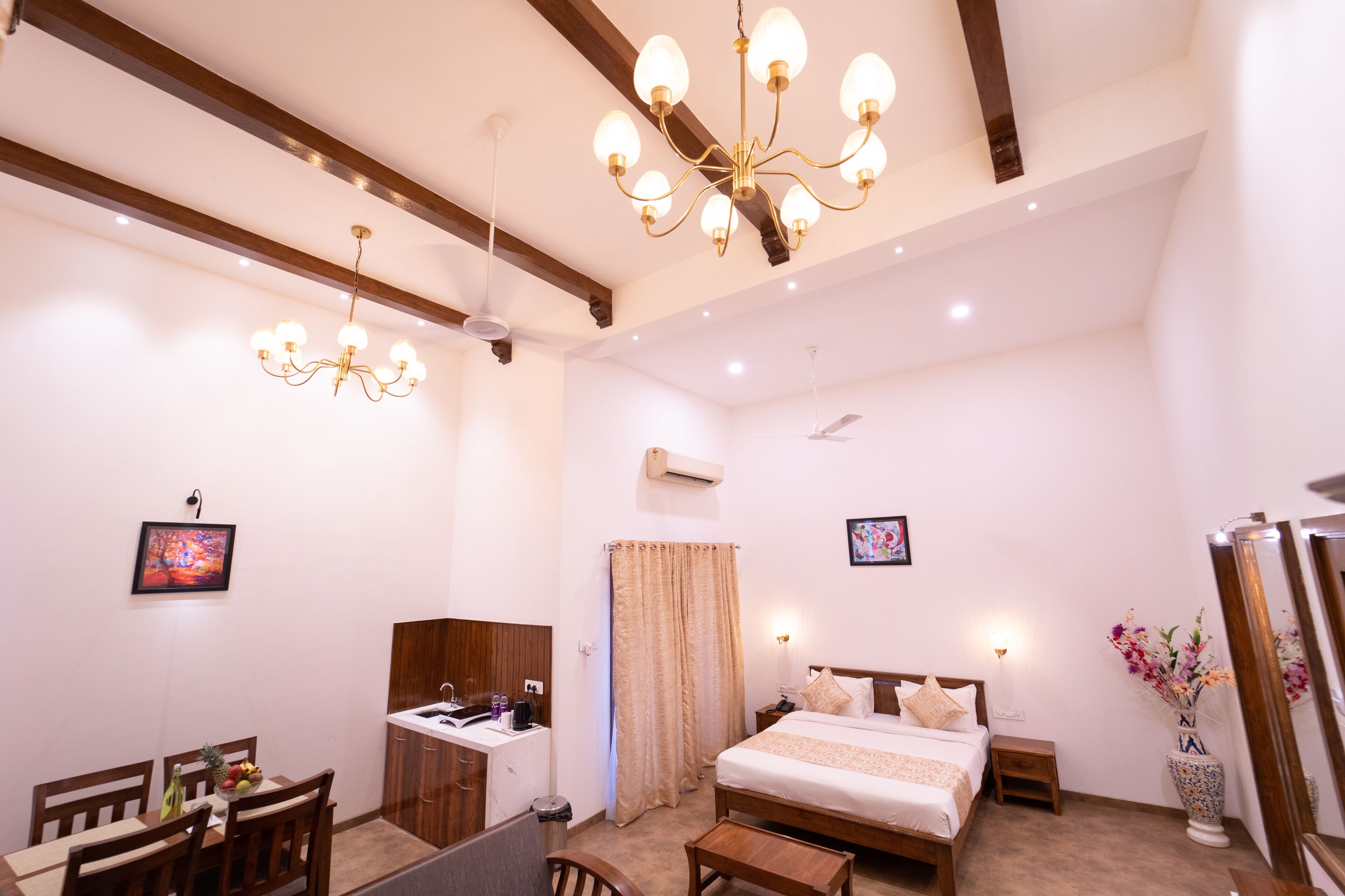A hotel room with a bed, bedside table, nightstand, and a desk with a chair. There is a chandelier, a window with curtains, and a vase with flowers.