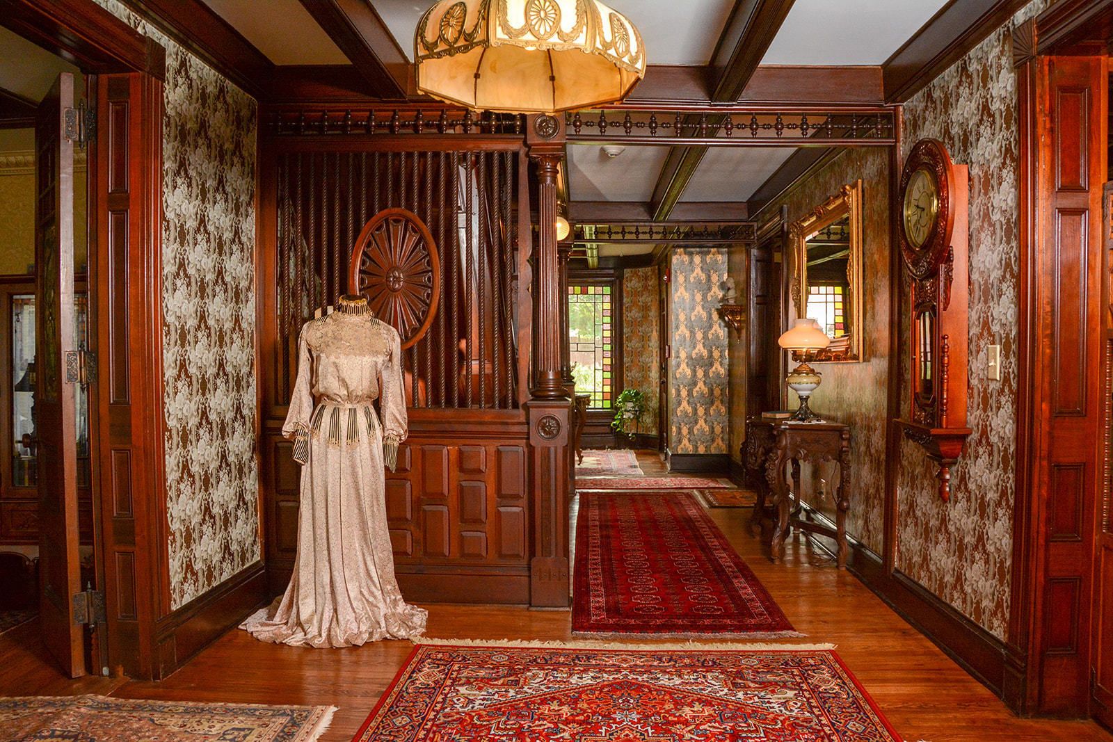 A grand hallway with a vintage dress on display, wooden paneling, and a red carpet.