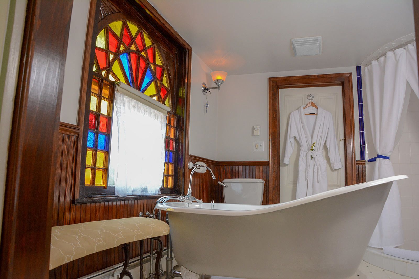 A bathroom with a freestanding bathtub, a wooden vanity, a stained glass window, and a robe hanging on the door.