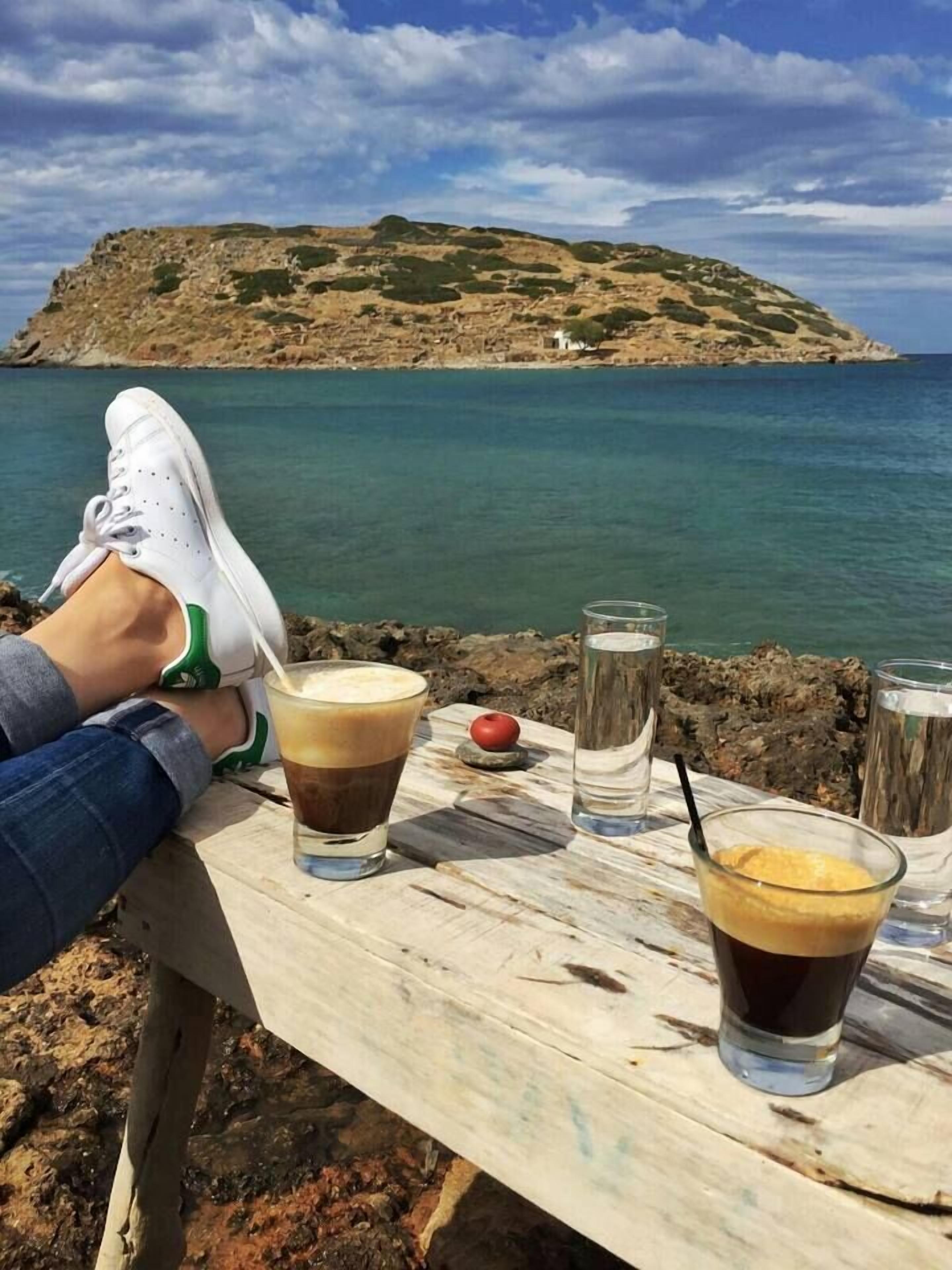 A wooden table with drinks and a person sitting with their feet up, overlooking a coastal landscape with a hill and sea.