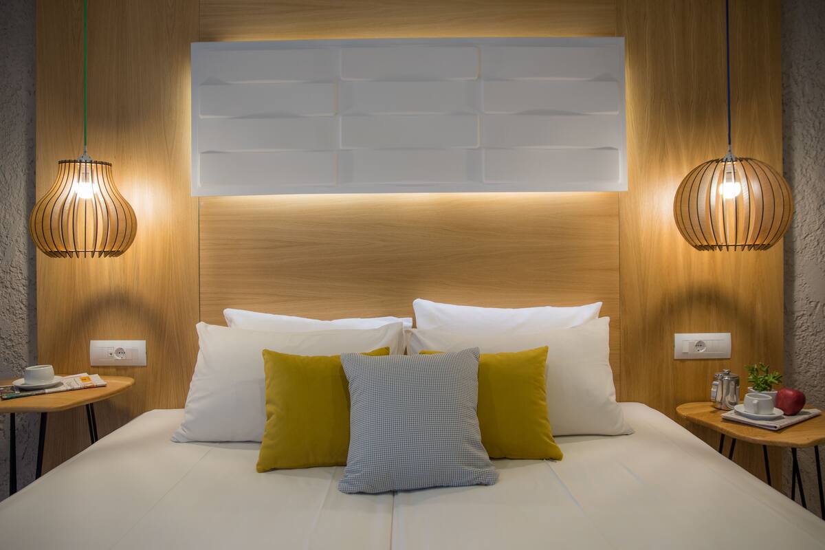 A neatly made bed with white and yellow pillows, two bedside tables with a cup and a book, and two hanging pendant lights.