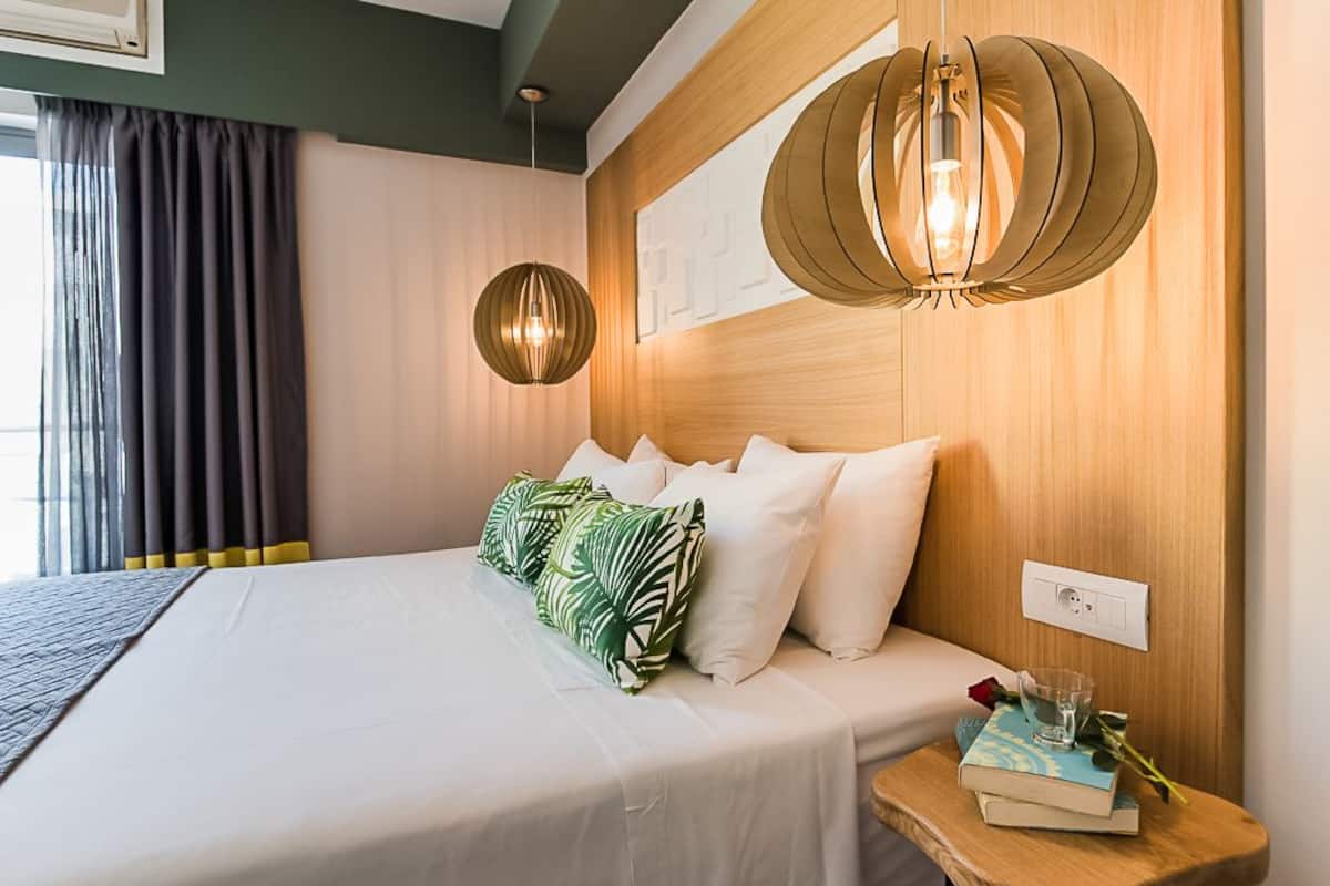A neatly made bed with white linens and green patterned pillows, a bedside table with a book and a glass, and a modern pendant light fixture.